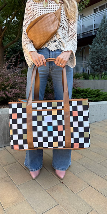 A person wearing a crocheted top, jeans, and pink heels holds a large fabric tote bag with a checkered pattern and brown handles. The person is also wearing a brown fanny pack around their waist. The tote's pattern is black and white with colored squares.
